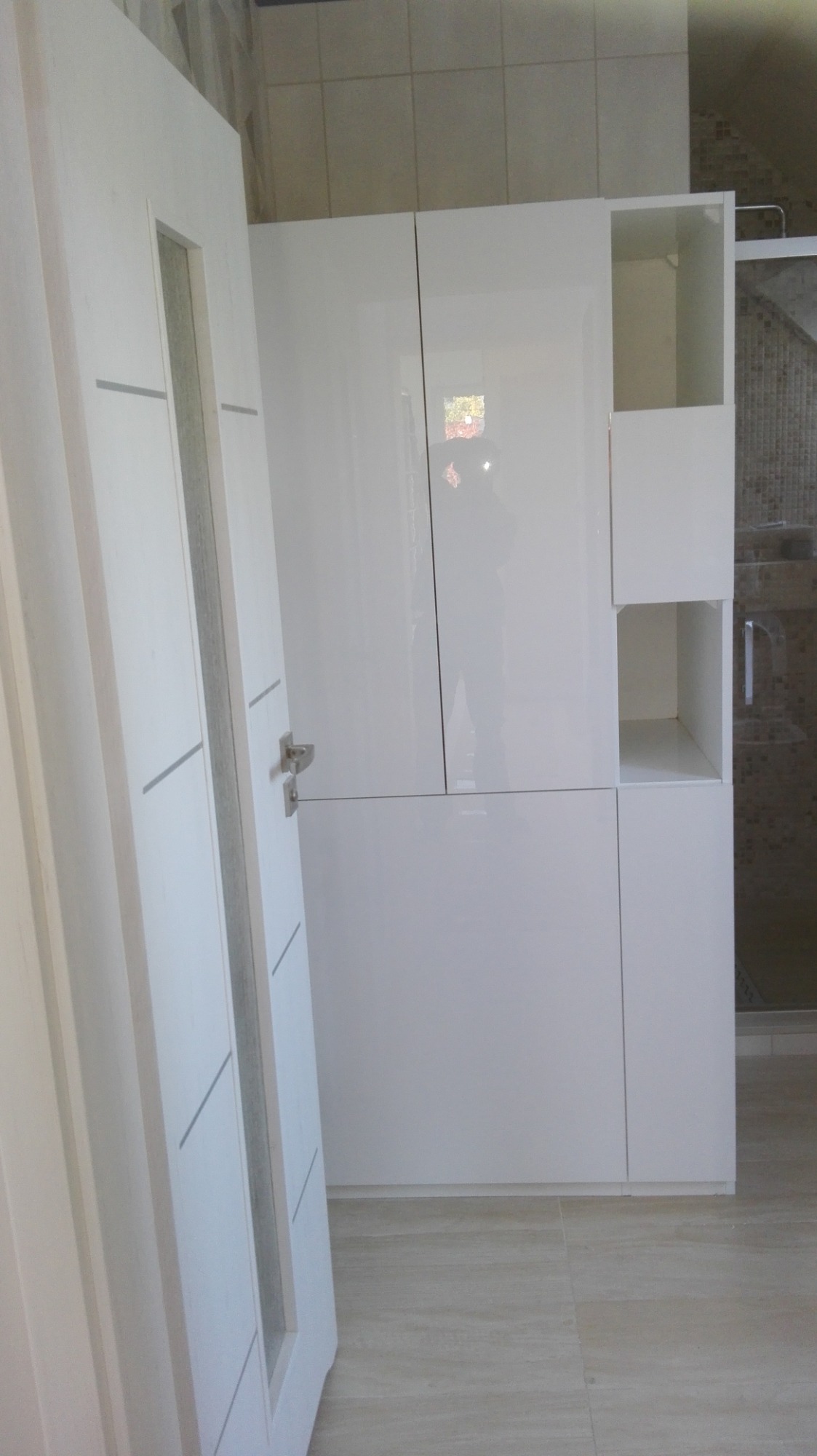 Biała szafa z otwartymi półkami w łazience, widoczna obok biała rama drzwi z szybą, płytki na podłodze i ścianach.