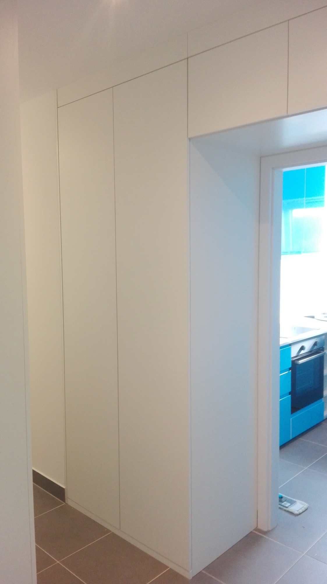 Biała zabudowa szaf w korytarzu, widok na fragment niebieskiej zabudowy kuchennej przez otwarte drzwi.