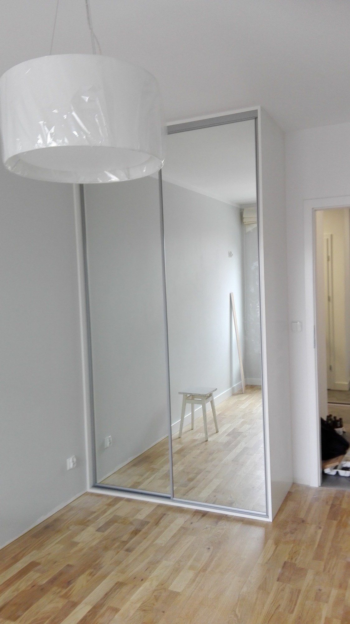 Minimalistyczna biała szafa z przesuwnymi lustrzanymi drzwiami, widoczna w jasnym pokoju z drewnianą podłogą, lampa w folii ochronnej zwisa z sufitu, w odbiciu krzesło i listwa przypodłogowa.