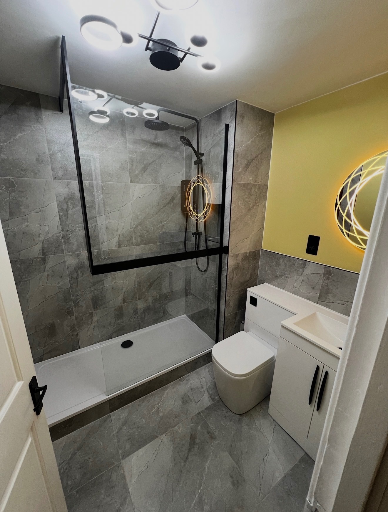 Nowoczesna łazienka z kabiną typu walk-in, szarymi płytkami imitującymi marmur i okrągłym lustrem z podświetleniem LED. Żółta ściana dodaje akcentu kolorystycznego.