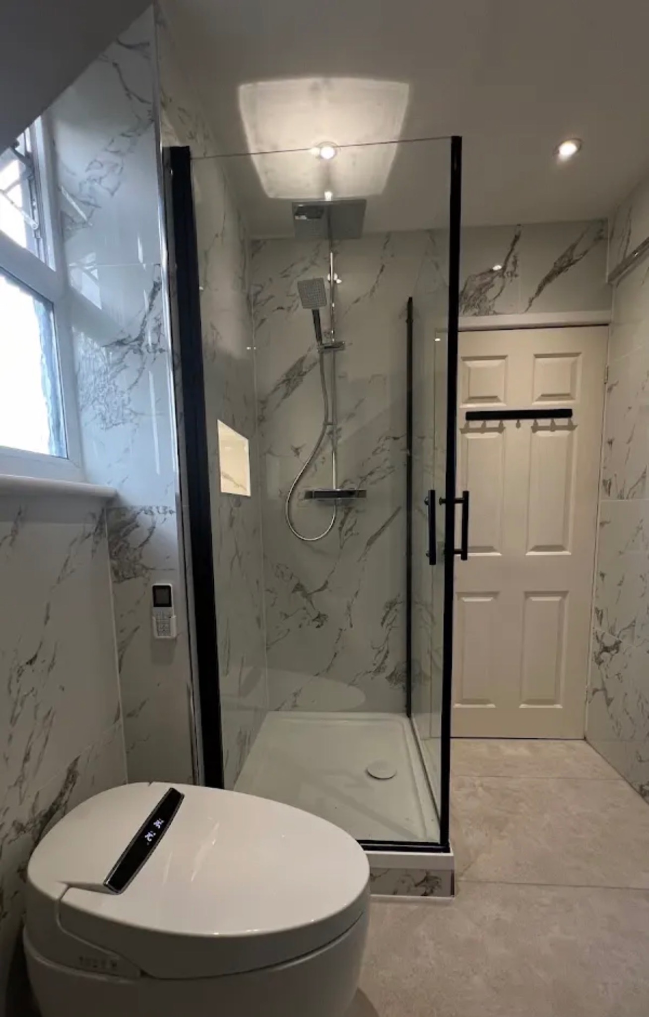 Nowoczesna łazienka z kabiną prysznicową typu walk-in, wykończona płytkami imitującymi marmur. Widoczny inteligentny sedes i drzwi.