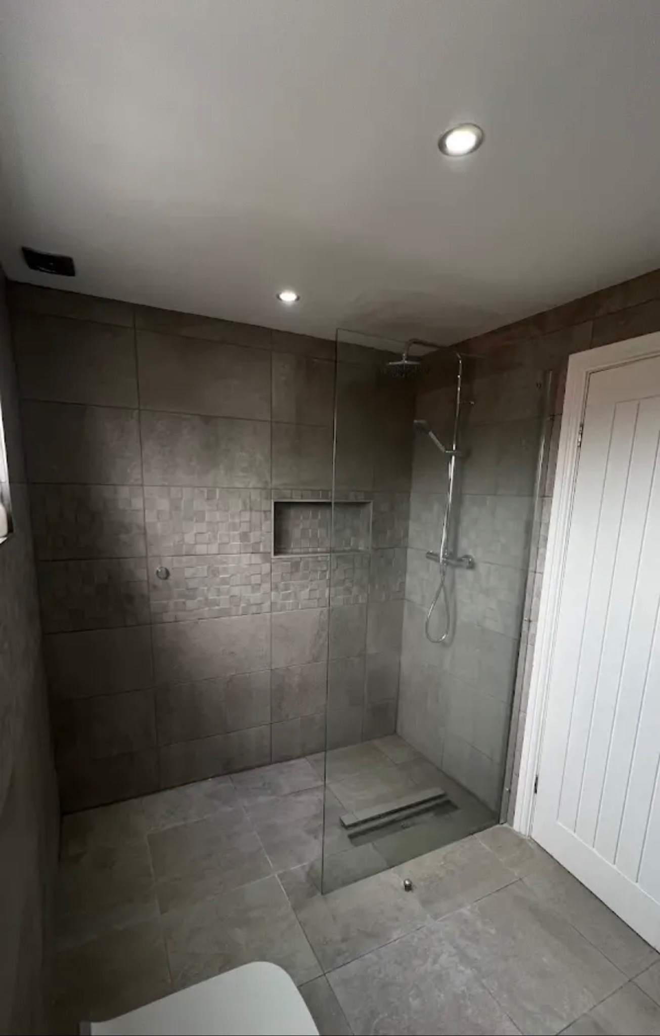 Nowoczesna łazienka z kabiną typu walk-in, wykończona szarymi płytkami w różnych odcieniach i wzorach. Widoczny odpływ liniowy i wnęka w ścianie.