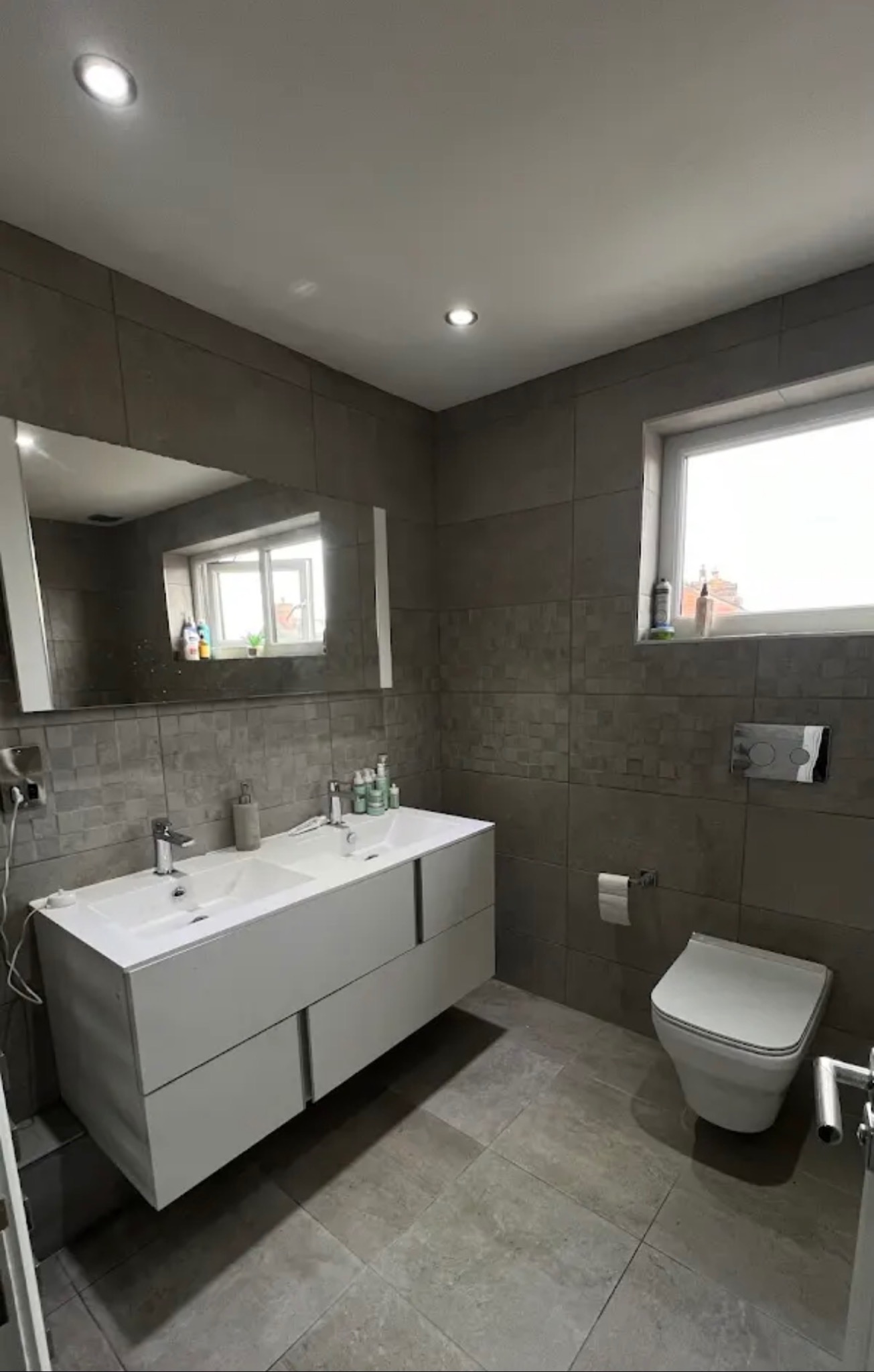 Nowoczesna łazienka w szarościach z podwójną umywalką na szafce, toaletą i oknem. Płytki imitujące kamień na ścianach i podłodze. Minimalistyczny design.