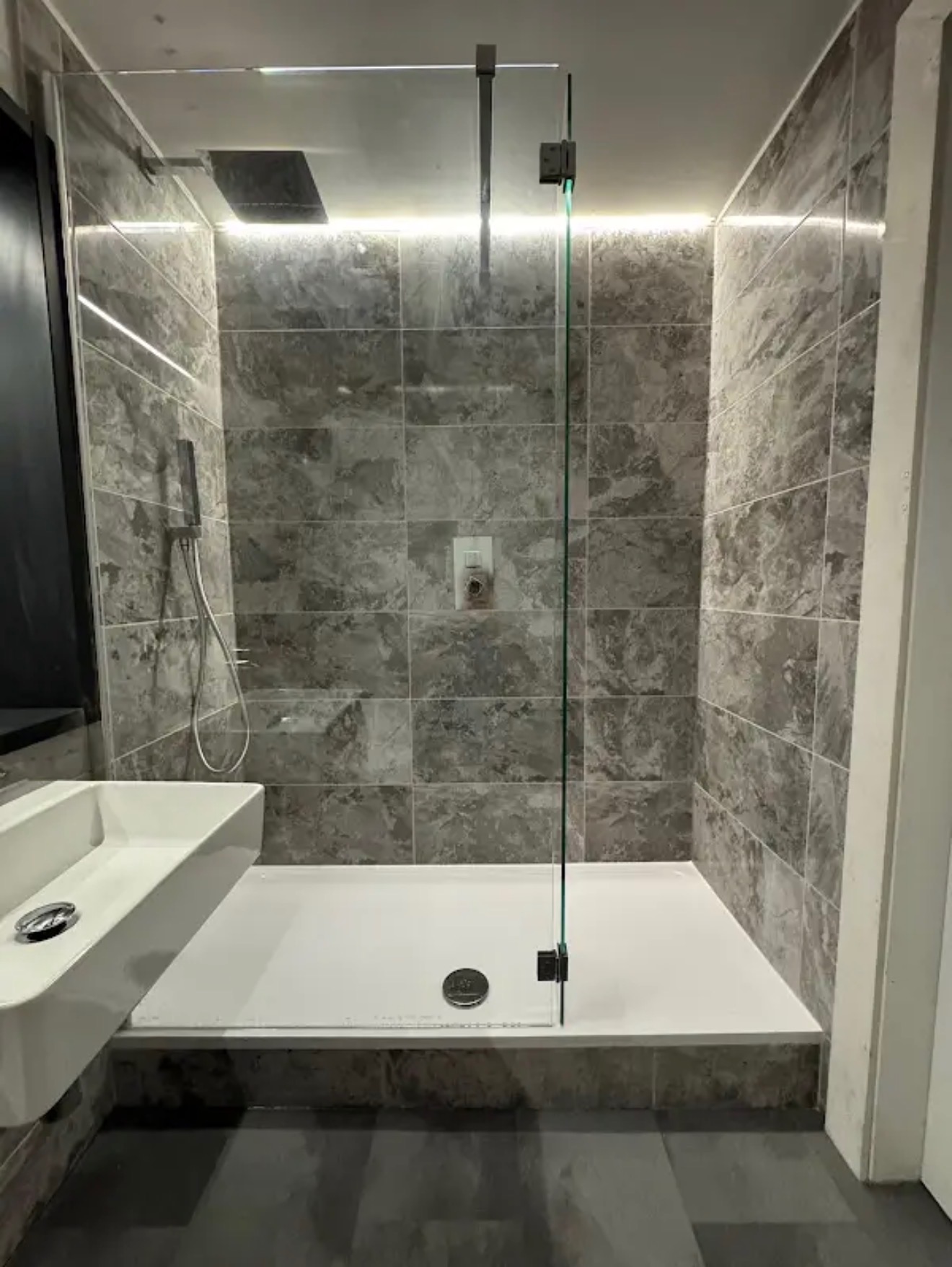 Nowoczesna łazienka z kabiną prysznicową typu walk-in, wykończona szarymi płytkami imitującymi marmur, z podświetleniem LED i białą umywalką.