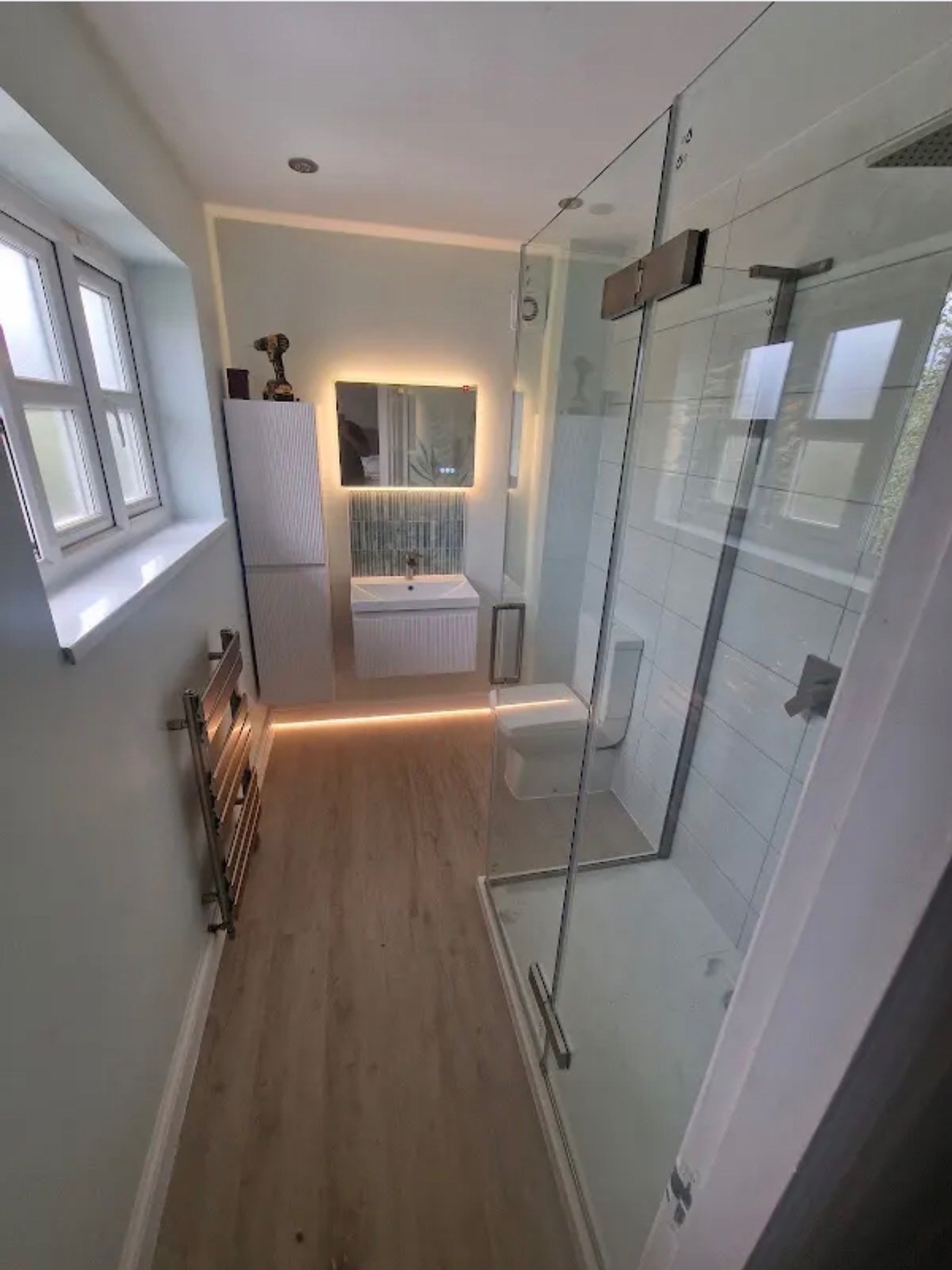 Nowoczesna łazienka z kabiną prysznicową ze szkła, podświetlanym lustrem, białymi meblami i drewnianą podłogą. Widoczny grzejnik drabinkowy i narzędzie na szafce.