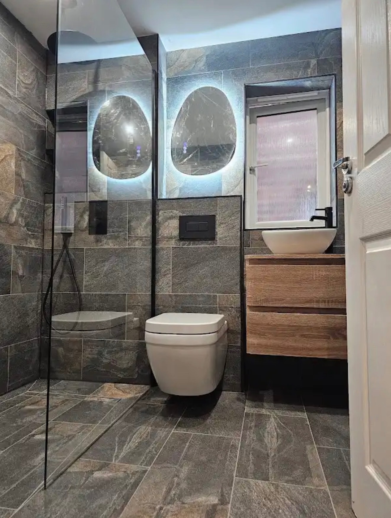 Nowoczesna łazienka z szarymi płytkami imitującymi kamień, podświetlanymi lustrami o nieregularnym kształcie i wiszącą miską WC. Kabina prysznicowa ze szkła.