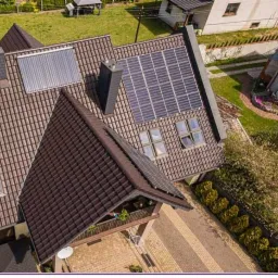 Widok z góry na dom z brązowym dachem, na którym zamontowano panele słoneczne i kolektor słoneczny.