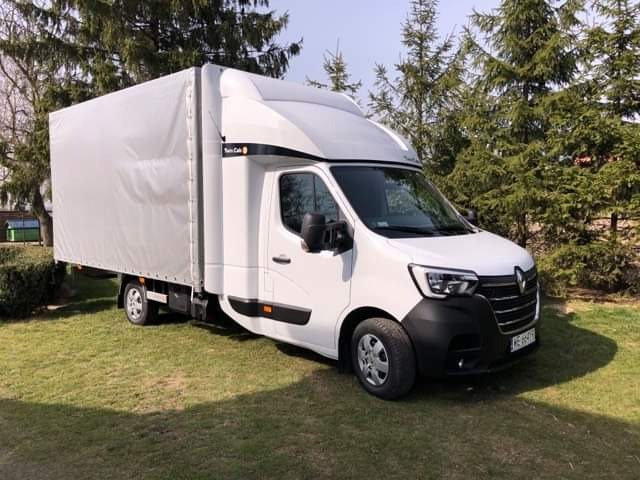 Biały samochód dostawczy z plandeką marki Renault Master na trawniku, widok z boku, tablica rejestracyjna widoczna.