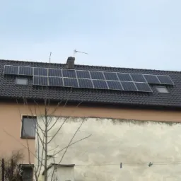 Panele słoneczne na ciemnym, skośnym dachu domu jednorodzinnego z oknami dachowymi, widoczne z poziomu ulicy w pochmurny dzień.