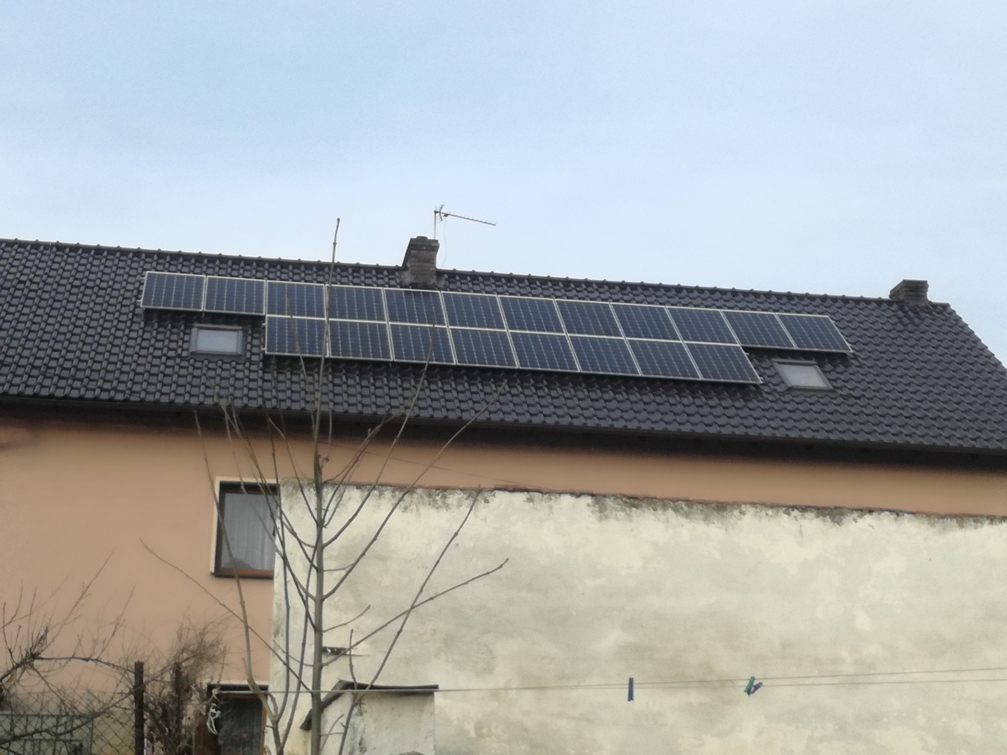 Panele słoneczne na ciemnym, skośnym dachu domu jednorodzinnego z oknami dachowymi, widoczne z poziomu ulicy w pochmurny dzień.