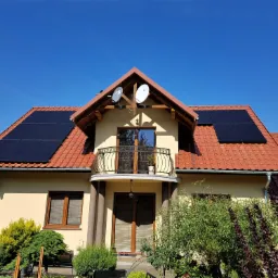 Dom jednorodzinny z panelami fotowoltaicznymi na dachu z czerwonej dachówki, dwie anteny satelitarne na szczycie dachu, jasna elewacja budynku i zadbany ogród.