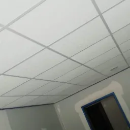 Kwadratowa, biała konstrukcja sufitowa z widoczną siatką profili, świeżo zamontowana w pomieszczeniu w trakcie prac wykończeniowych; ściany w szarym kolorze z widocznymi śladami szpachlowania...