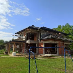 Budowa domu w stanie surowym otwartym, widoczne rusztowania, częściowo pokryty dach z dachówki ceramicznej, pracownik na dachu, niebieska bramka piłkarska w pierwszym planie.