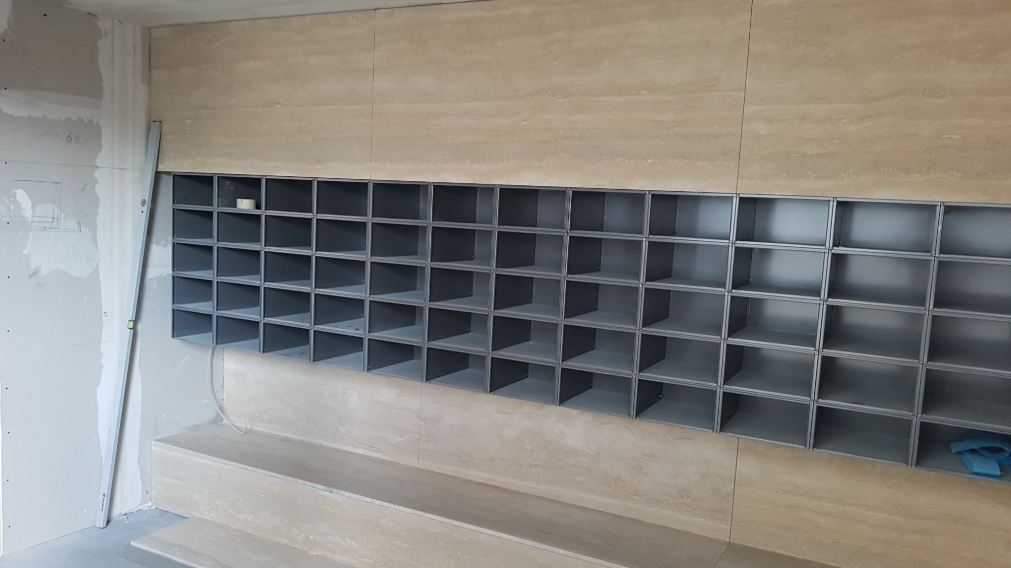 Ściana z trawertynu z wnękami w trakcie montażu, poziomica przy ścianie z płyt gipsowych, widoczne okablowanie i taśma maskująca.