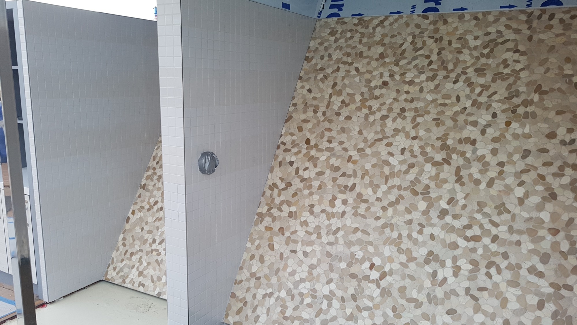 Fragment łazienki w trakcie wykańczania: białe kwadratowe płytki na ścianie z zainstalowanym zaworem obok ściany wyłożonej mozaiką z drobnych, beżowych kamyczków.