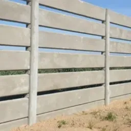 Długi, szary płot z prefabrykowanych element&oacute;w betonowych z poziomymi szczelinami, widok z boku, zielona trawa u podn&oacute;ża, błękitne niebo w tle.