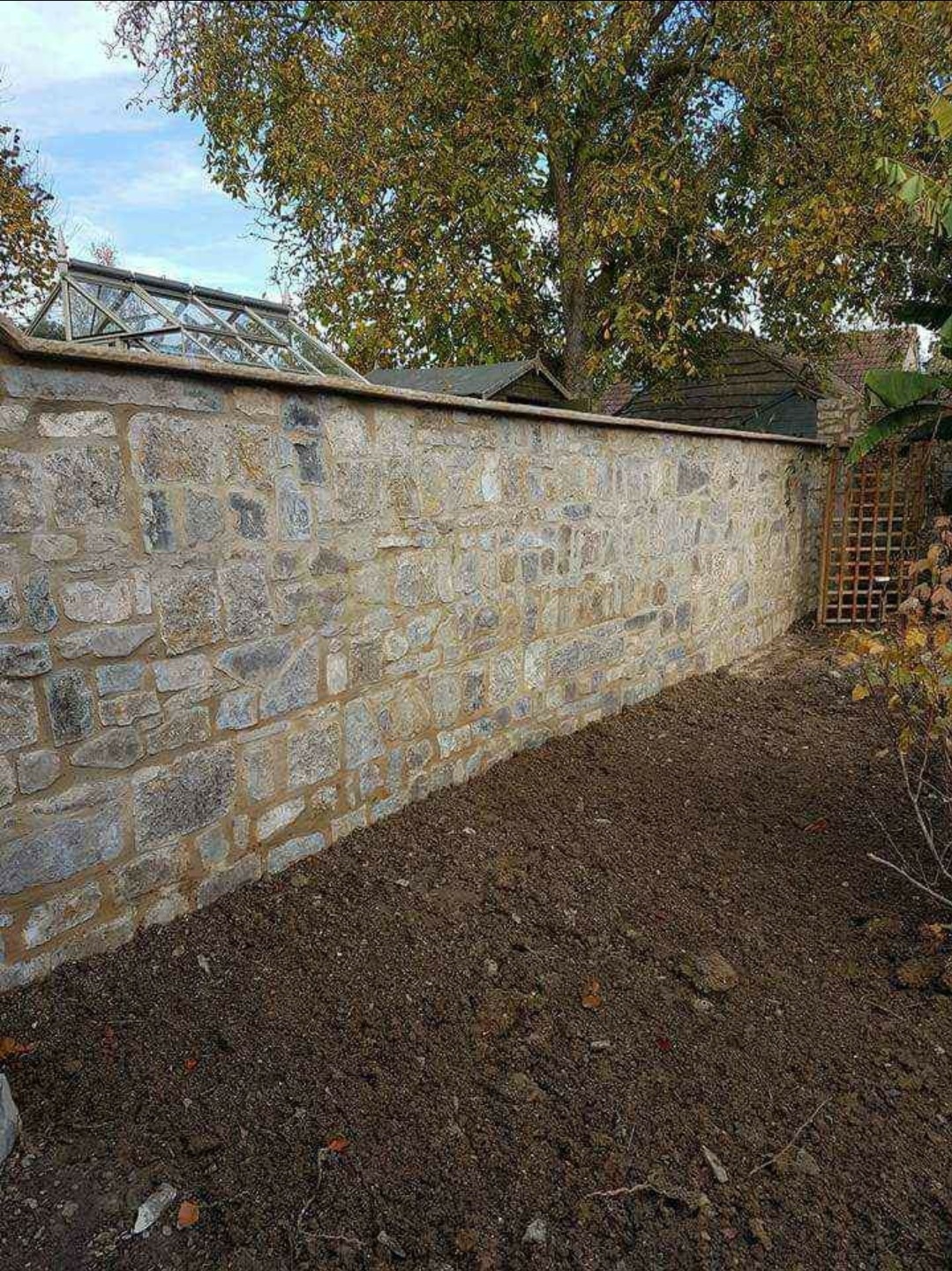 Masywny mur z nieregularnych kamieni o szarej barwie, z widocznymi spoinami, zakończony płaskim daszkiem, przylegający do ziemi i otoczony roślinnością.