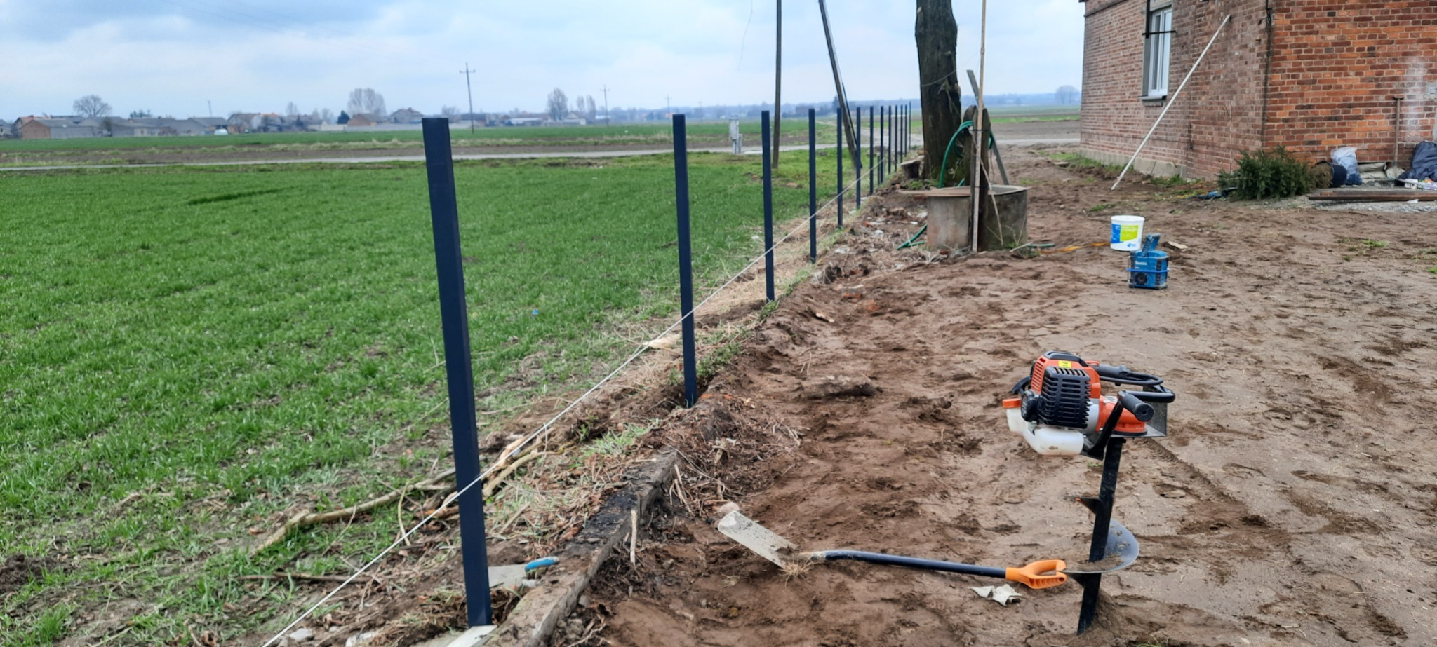 Wkopane słupki ogrodzeniowe z naciągniętym sznurkiem wyznaczającym linię, w tle zielone pole i zabudowania wiejskie, na pierwszym planie wiertnica spalinowa do gruntu i łopata.