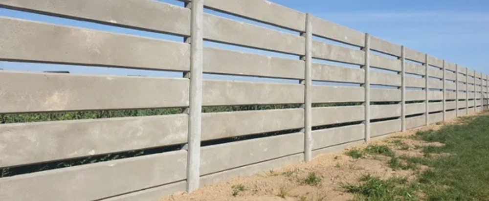 Długi, szary płot z prefabrykowanych elementów betonowych z poziomymi szczelinami, widok z boku, zielona trawa u podnóża, błękitne niebo w tle.