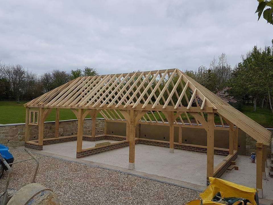 Drewniana konstrukcja z zadaszeniem w trakcie budowy, widoczne elementy więźby dachowej, murowane fragmenty ścian, krajobraz w tle.