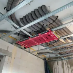 Montaż systemu rekuperacji z szarymi rurami wentylacyjnymi podwieszonymi do metalowej konstrukcji sufitu podwieszanego, widoczny moduł rekuperatora w czerwonej obudowie Red Snake.