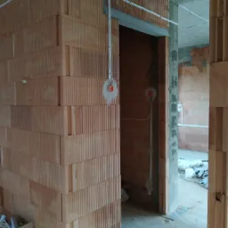 Wnętrze w trakcie budowy z widocznymi ścianami z cegieł ceramicznych, przewodami elektrycznymi prowadzonymi po powierzchni ścian i przygotowanymi otworami pod puszki elektryczne.