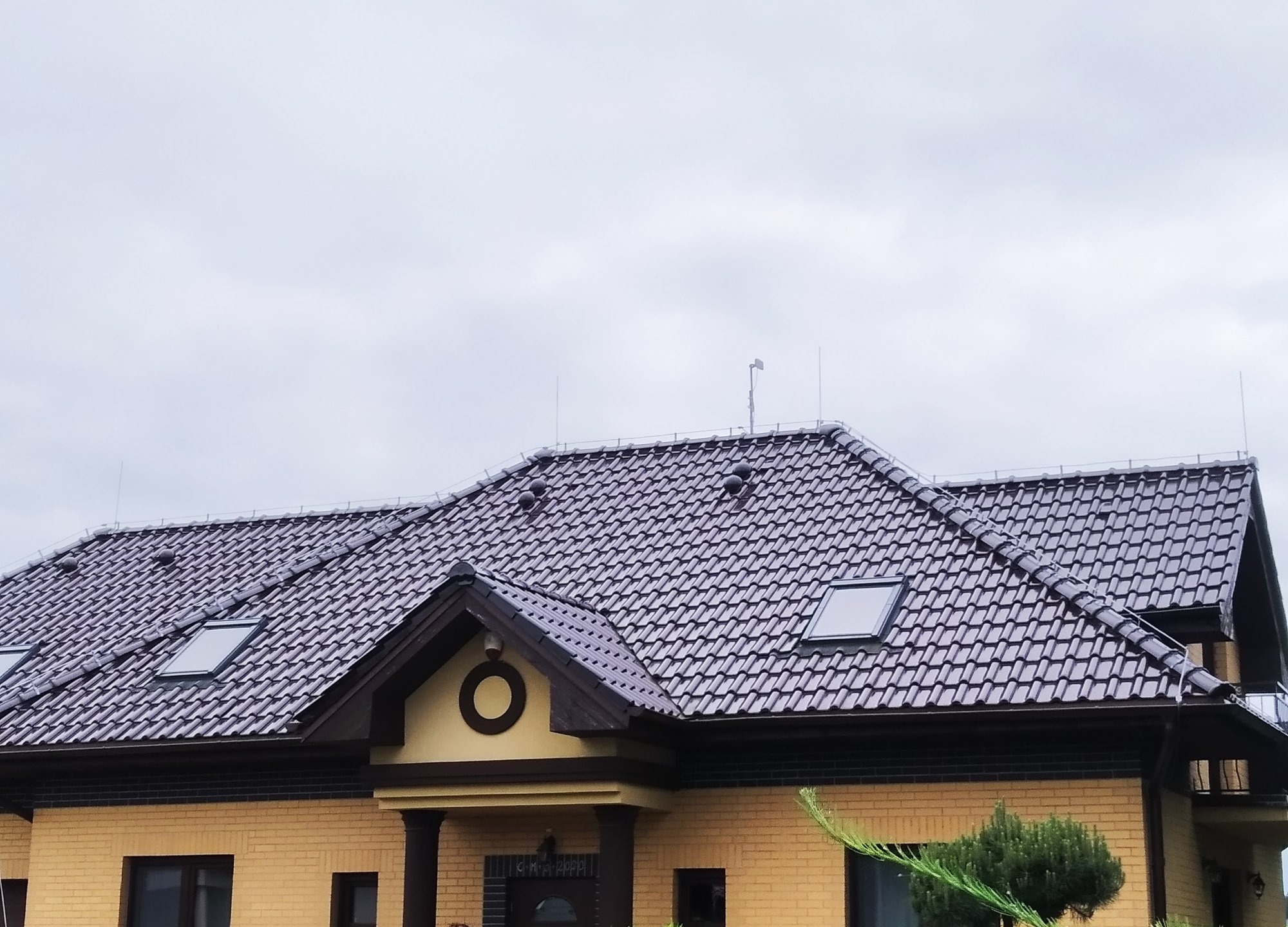 Dwukondygnacyjny dom z żółtej cegły z brązowym dachem z dachówki ceramicznej, widoczne okna dachowe i piorunochrony na krawędzi dachu.