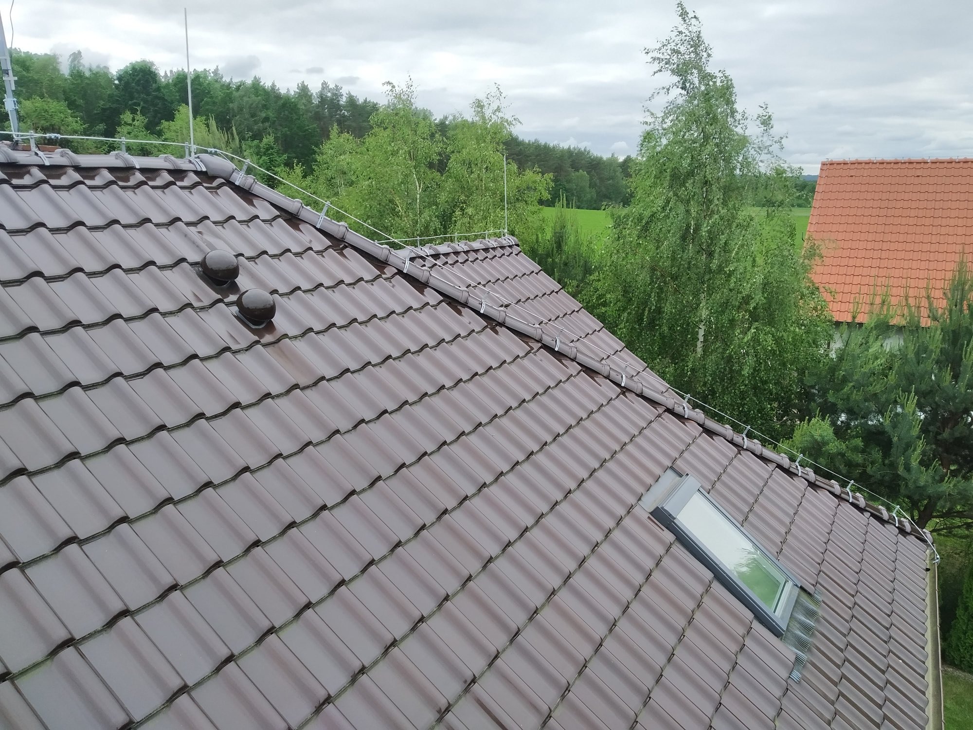 Ciemnobrązowy dach ceramiczny z oknem dachowym i instalacją odgromową, widok z góry na tle zieleni.