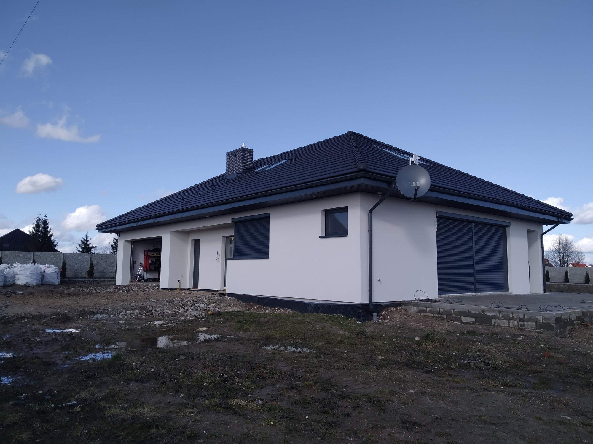 Niedawno wybudowany, jednopiętrowy dom z szarą dachówką i białymi ścianami, z roletami zewnętrznymi, widoczny z zewnątrz, z niezagospodarowanym terenem wokół.