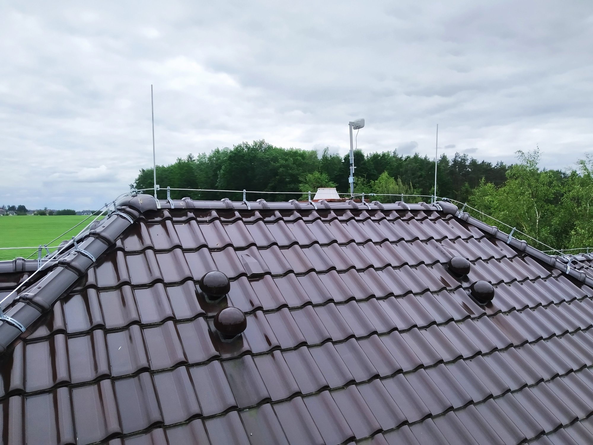 Brązowy dach z dachówki ceramicznej z zamontowaną instalacją odgromową i odpowietrznikami w Chojnicach, na tle pochmurnego nieba i zieleni drzew.