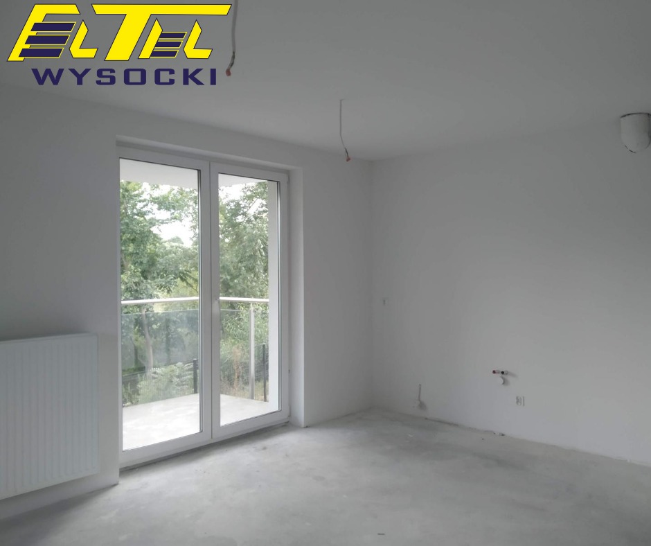 Surowe wnętrze nowo wybudowanego domu z białymi ścianami, betonową podłogą, grzejnikiem i oknem balkonowym z widokiem na zieleń, widoczne wystające przewody elektryczne ze ściany i logo firmy...