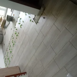 Układanie podłogi z płytek ceramicznych w odcieniach beżu, z użyciem zielonych krzyżyków dystansowych, widoczny kubeł z zaprawą i paczka płytek, ułożone pod kątem.