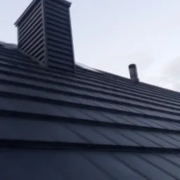 Ciemnoszary dach z blachy trapezowej z widocznym kominem wentylacyjnym i odpowietrznikiem kanalizacji na tle pochmurnego nieba.