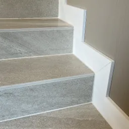 Wykończone schody z szarych płytek imitujących kamień, z białymi listwami przypodłogowymi i krawędziami schodów, na tle ściany w odcieniu beżu.