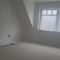 Poddasze po szpachlowaniu i malowaniu na biało, okno zabezpieczone folią malarską, widoczne ślady po gładzi na podłodze, przygotowane do dalszych prac wykończeniowych.