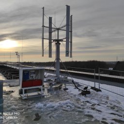 Hipar Sp. z o.o. - Pionowa turbina wiatrowa na dachu budynku pokrytym śniegiem i lodem, widoczne panele i skrzynka elektryczna, w tle zachodzące słońce.