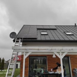 Hipar Sp. z o.o. - Montaż paneli słonecznych na dachu domu z metalową dachówką, widoczna drabina oparta o dach i satelita, pracownik przygotowujący się do instalacji na zewnątrz.