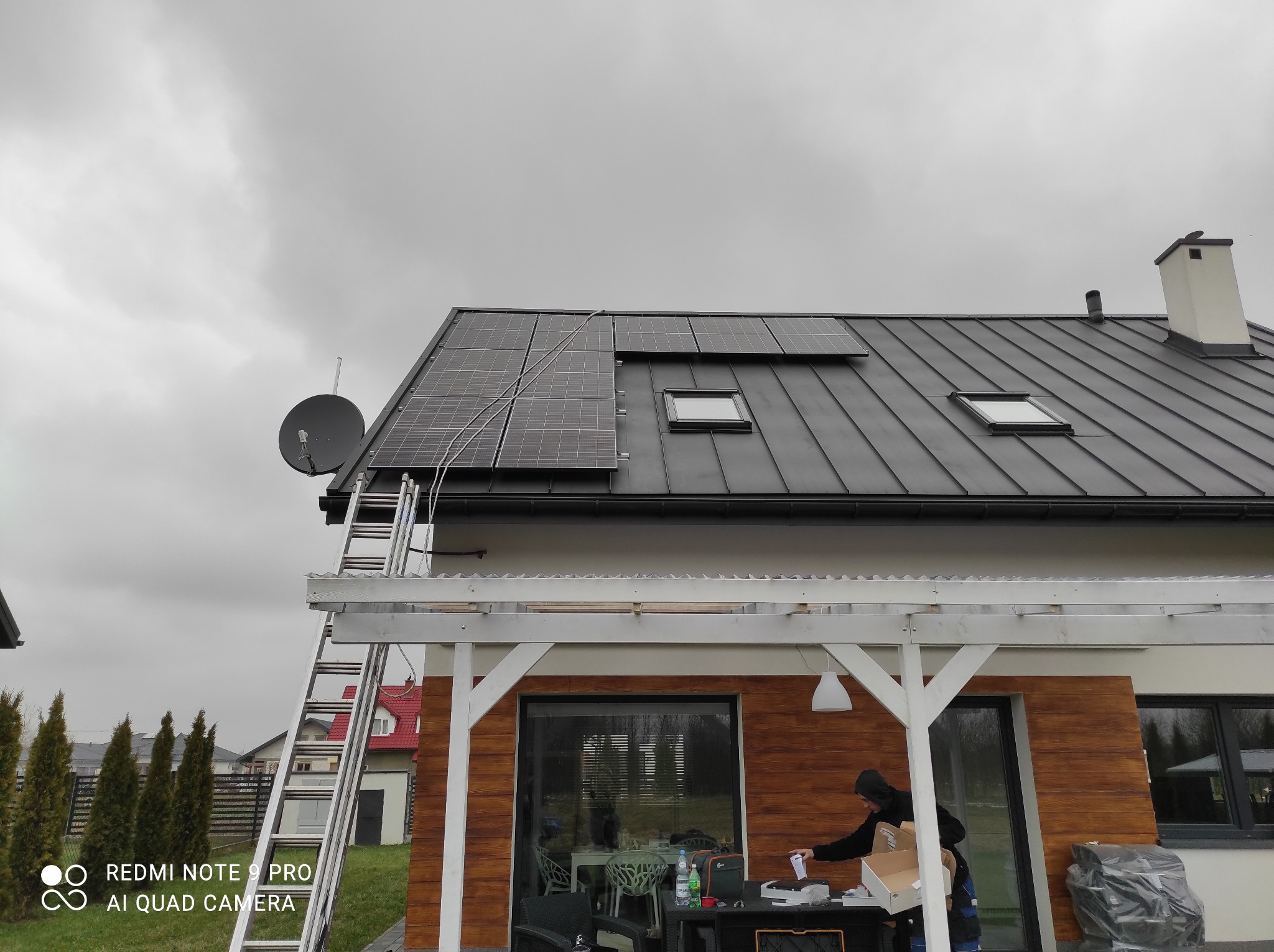 Montaż paneli słonecznych na dachu domu z metalową dachówką, widoczna drabina oparta o dach i satelita, pracownik przygotowujący się do instalacji na zewnątrz.