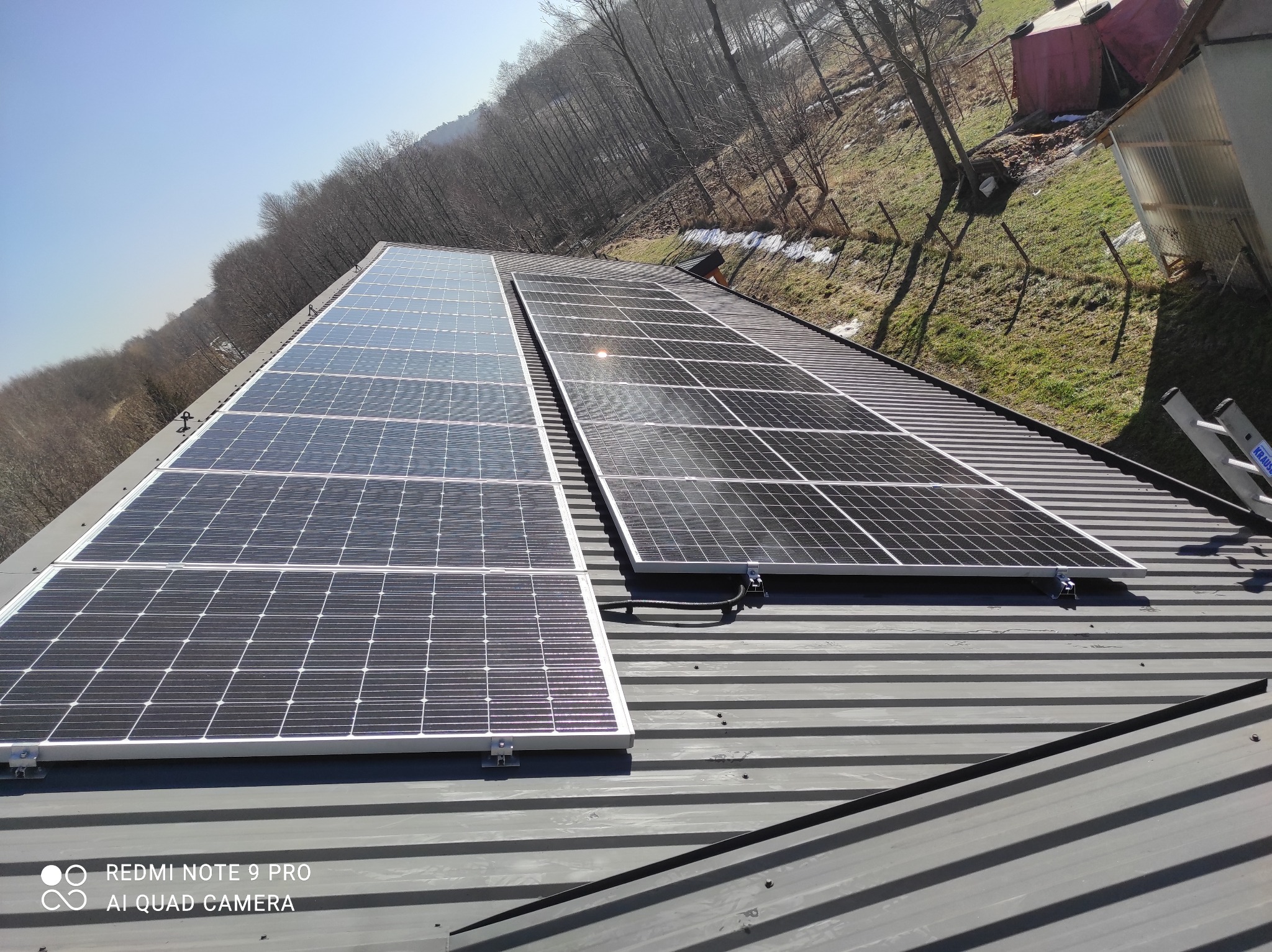 Panele słoneczne zamontowane na szarym, trapezowym dachu, z widocznym krajobrazem drzew bez liści i fragmentami zieleni w tle. Na prawo od paneli widoczna drabina.