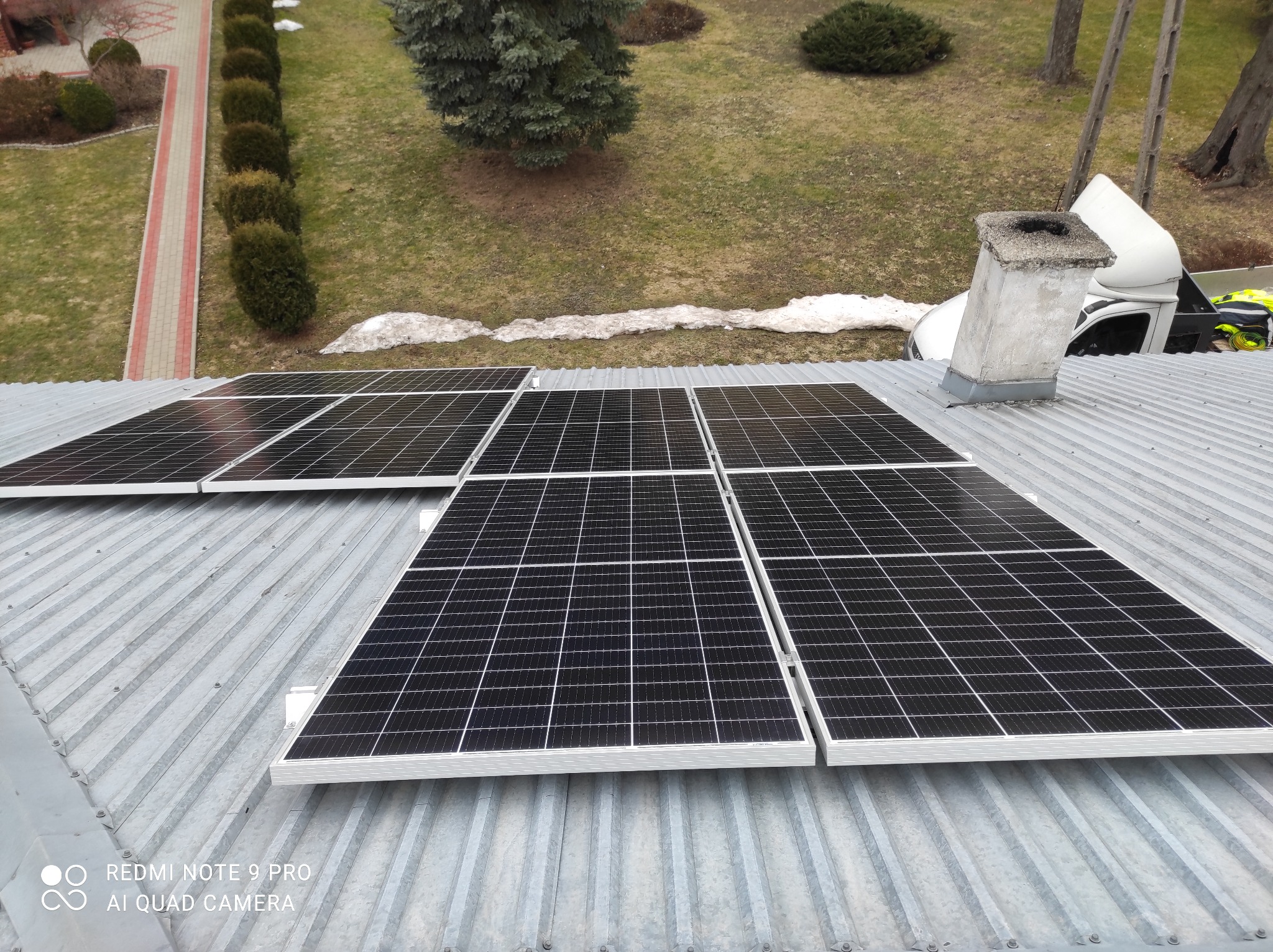 Panele słoneczne zamontowane na blaszanym dachu, widok z góry na ogród z resztkami śniegu i komin z samochodem dostawczym w tle.