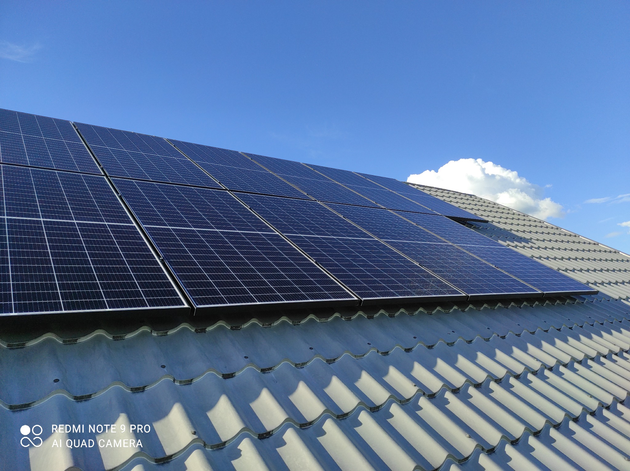 Panele słoneczne na szarym dachu z ceramicznej dachówki, widoczne na tle błękitnego nieba z pojedynczą białą chmurą.