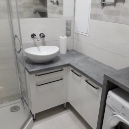 BBud - Nowoczesna łazienka z białą umywalką nablatową, szarą armaturą i blatem. W tle widoczne lustro i kabina prysznicowa. Minimalistyczny design.