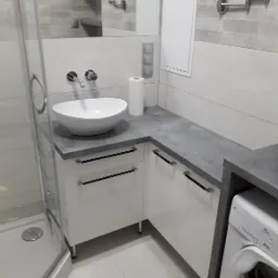 Nowoczesna łazienka z białą umywalką nablatową, szarą armaturą i blatem. W tle widoczne lustro i kabina prysznicowa. Minimalistyczny design.