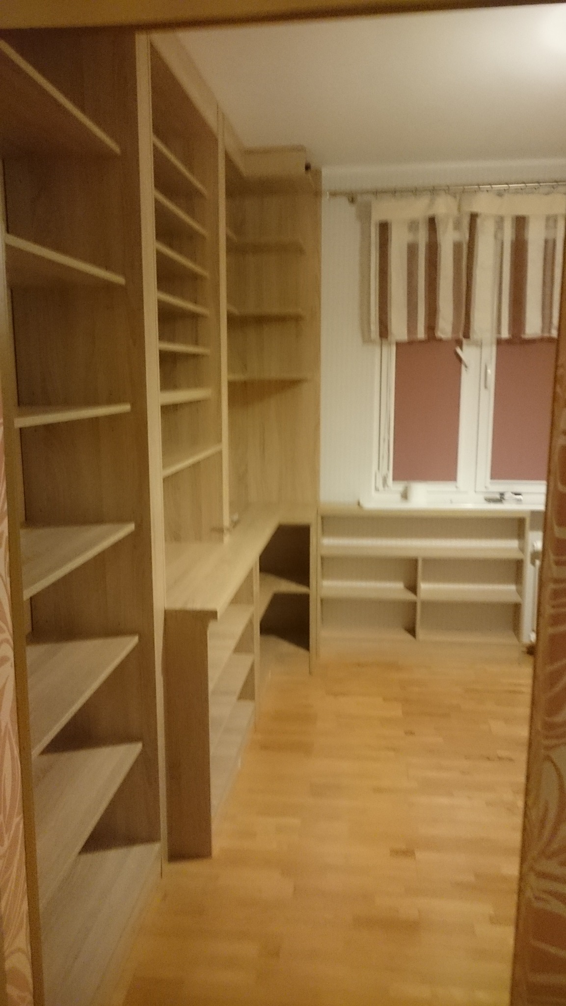 Jasne, drewniane regały z półkami, zabudowa narożna z blatem biurka, widok na okno z zasłonami w paski, parkiet na podłodze.