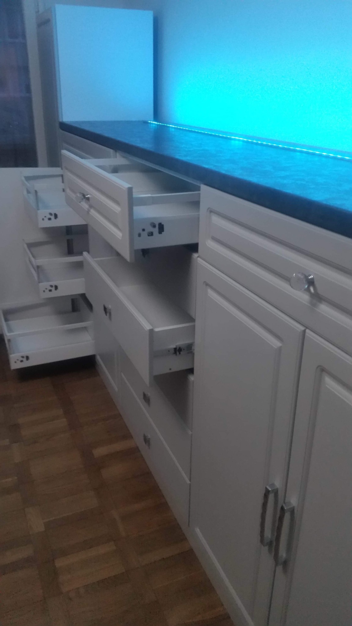 Białe szafki kuchenne z otwartymi szufladami, podświetlane niebieskim światłem LED, drewniana podłoga parkietowa, srebrne uchwyty i kryształowe gałki.