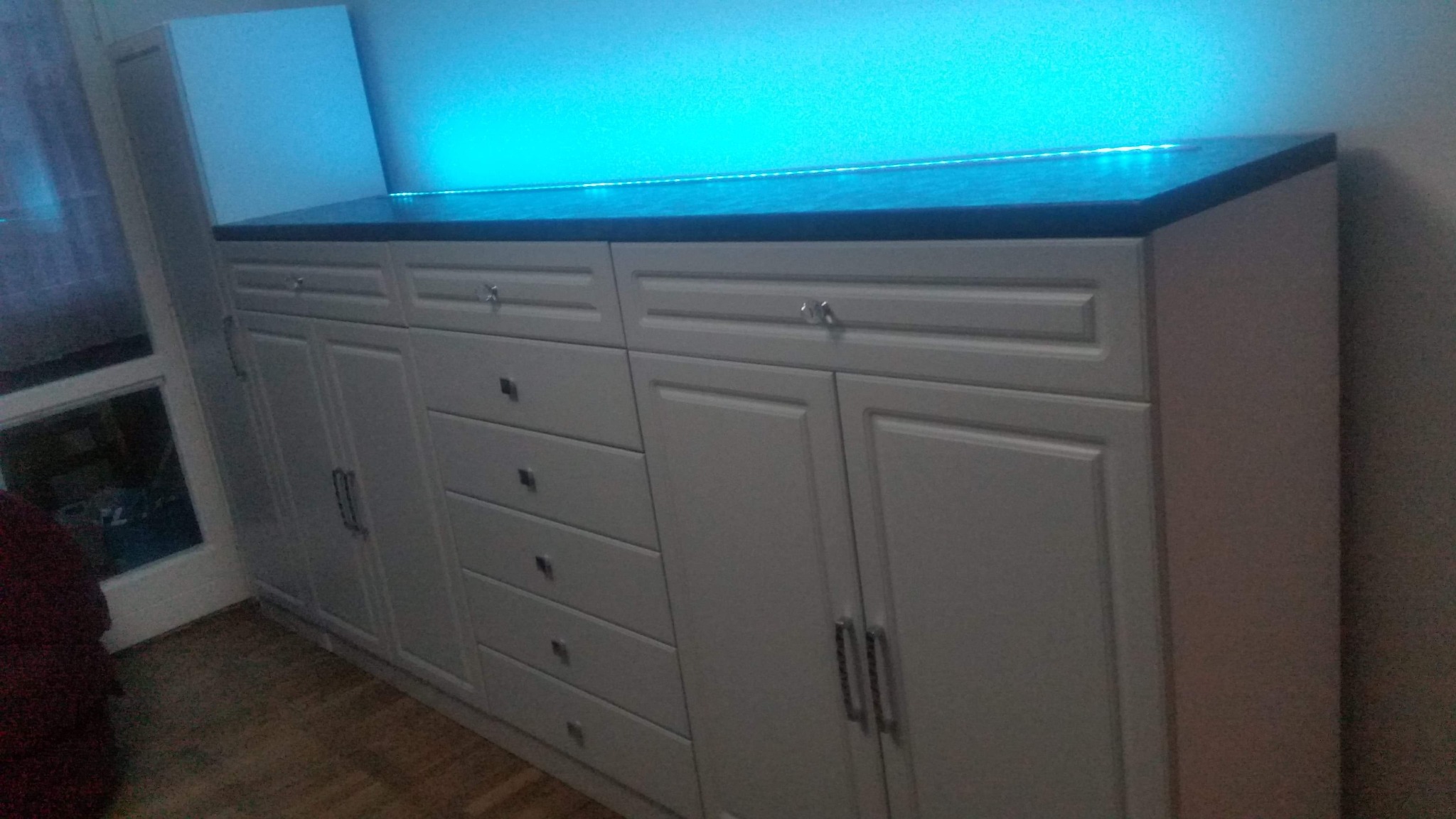 Biała zabudowa meblowa z szufladami i szafkami, czarny blat, podświetlenie LED. Widoczne srebrne uchwyty i dekoracyjne gałki. Ujęcie pod kątem z boku.