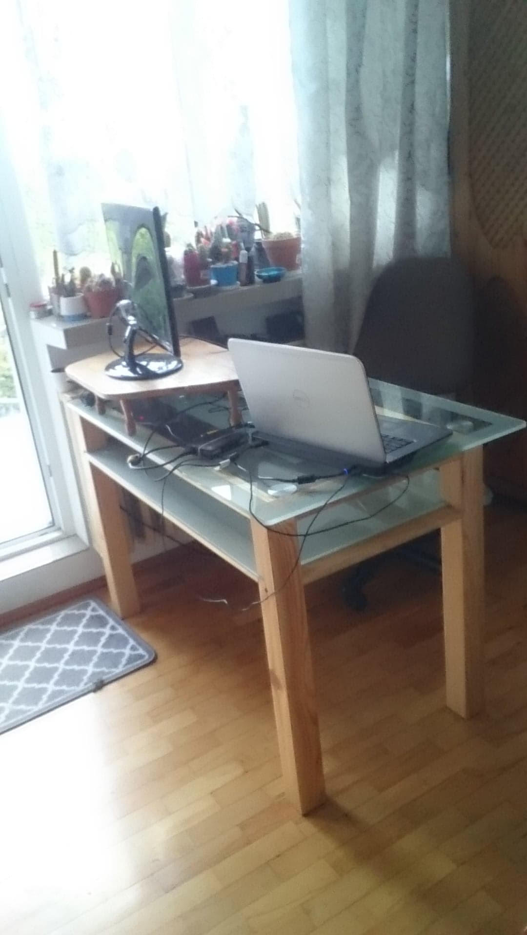 Szklany stolik z laptopem i monitorem w Szczecinie, ustawiony przy oknie z zasłonami. Widoczne drewniane nogi i półki stolika, dywanik pod spodem.