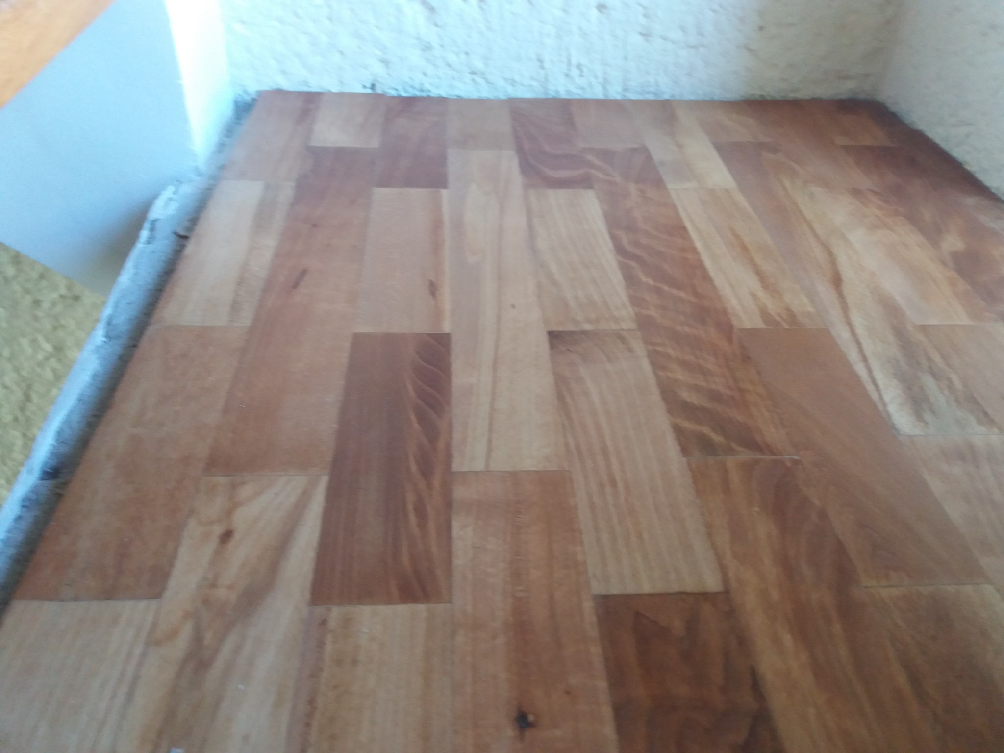 Ułożona, lakierowana podłoga drewniana z parkietu w odcieniach brązu, z widocznymi słojami, przy ścianie wykończonej szarym cokołem.