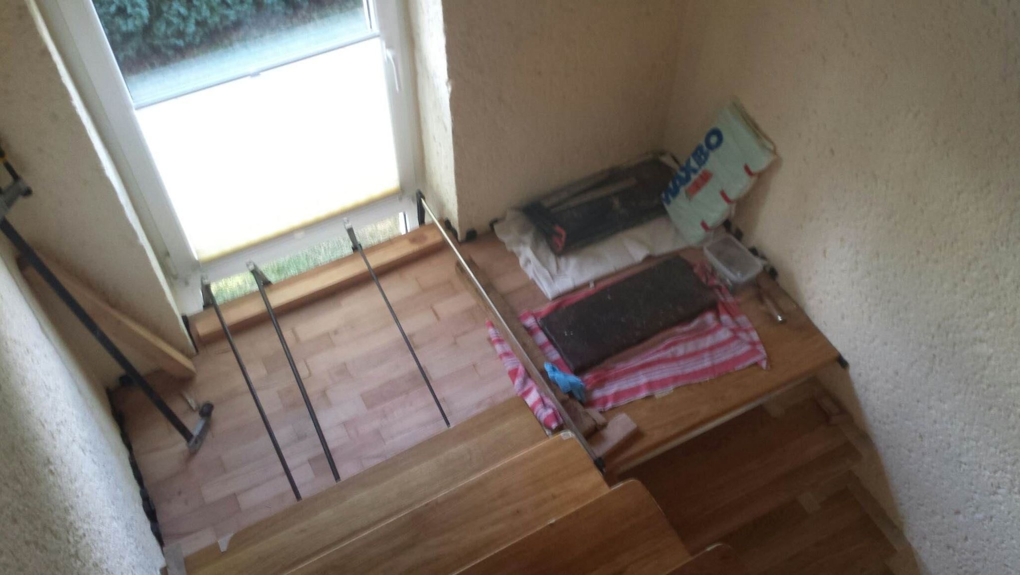 Montaż drewnianych schodów z balustradą przy oknie. Widoczne narzędzia i materiały budowlane, etap prac wykończeniowych w jasnym wnętrzu.