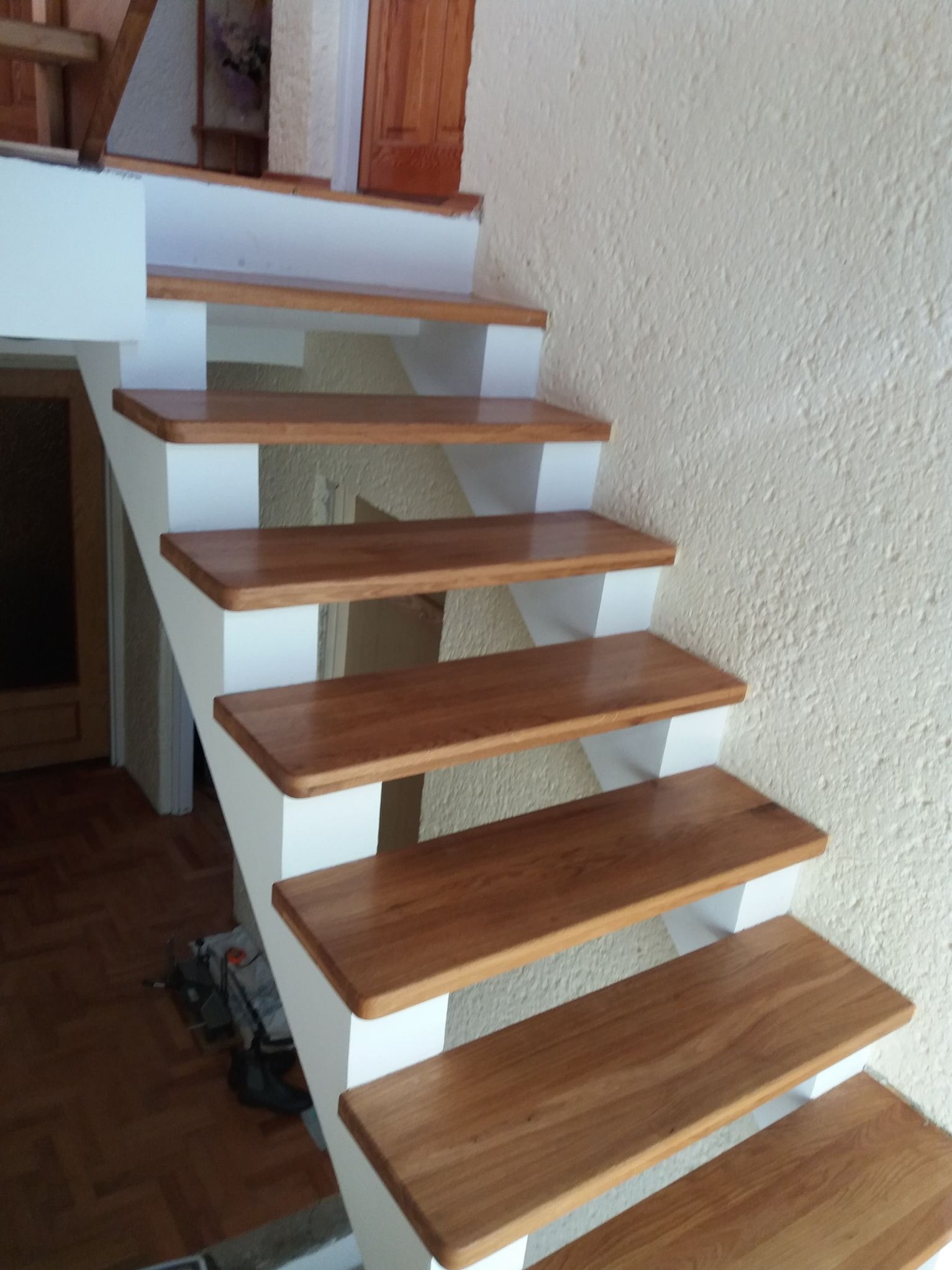 Drewniane schody z białymi wspornikami na tle beżowej ściany o nierównej fakturze. Widok z dołu, ukazujący detale wykończenia i solidną konstrukcję.